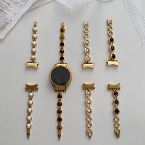 Könnte beinhalten: Eine goldfarbene Smartwatch mit rundem Zifferblatt und sieben austauschbaren Armbändern. Die Armbänder bestehen aus goldfarbenen Metallgliedern, die mit weißen runden Perlen oder braunen Tigeraugen-Steinen in verschiedenen Mustern verziert sind.