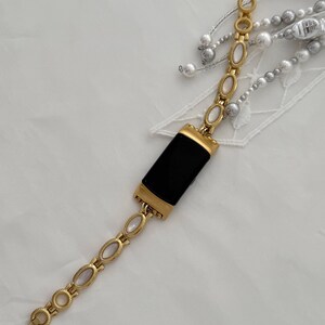 Op de afbeelding: Een fitnesstracker met een rechthoekig zwart scherm en een goudkleurige metalen behuizing. De band bestaat uit goudkleurige ovale schakels, elk met een centrale witte inleg, verbonden door gestructureerde randen in een touwachtig patroon.