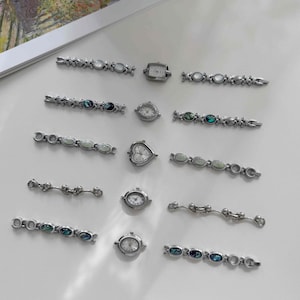 Peut inclure: Une collection de cinq cadrans de montre argentés et dix bracelets assortis disposés par paires. Les cadrans présentent des formes rectangulaires, rondes et en cœur. Les bracelets comportent divers maillons décoratifs, dont des incrustations en nacre d'ormeau irisée et des accents ovales blancs lisses.