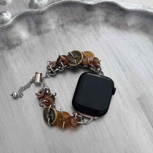 May include: A watch band with a unique design. The band features a series of brown buttons and a silver chain. The watch face is black. The band is attached to a silver watch case.