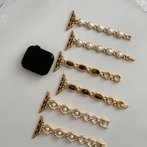 May include: A black smartwatch case and six gold-toned metal watch bands. Two bands feature white pearl-like beads, two feature brown oval tiger eye-style stones, and two feature gold-toned circular links with small white pearl-like accents.