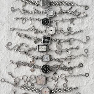 Könnte beinhalten: Eine Auswahl an silberfarbenen Uhren mit verschiedenen Zifferblattformen und dekorativen Anhängern. Die Uhren verfügen über verschiedene Armbandstile, darunter Kettenglieder und Perlendesigns. Die Zifferblätter sind weiß oder schwarz, mit römischen Ziffern oder einfachen Markierungen.