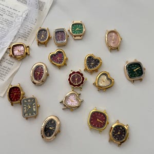 Pode incluir: Uma coleção de mostradores de relógio em tom dourado com diversas formas, incluindo corações, círculos, quadrados e retângulos. Os mostradores apresentam cores variadas como vermelho, verde, preto e rosa, alguns com algarismos romanos.