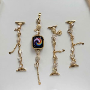 May include: Gold Apple Watch with a rainbow display, and several decorative bands. The bands are adorned with charms including stars, butterflies, and flowers, with dangling chains. The watch and bands are set against a white background.