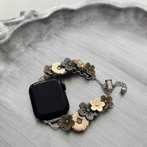 May include: A black smartwatch with a floral band. The band features flowers in gold, bronze, and gray tones, connected by a silver chain. The watch is displayed on a gray, textured surface.