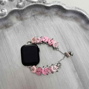 Pode incluir: Um smartwatch preto com uma pulseira decorativa. A pulseira é feita de pequenos botões rosa em várias tonalidades, incluindo lisos, listrados e brilhantes, conectados por elos de corrente prateados.