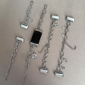 Op de afbeelding: Een collectie zilverkleurige metalen horlogebandjes en accessoires met schakels, karabijnsluitingen en kleine bedels zoals een zon, sterren en een maansikkel. Eén bandje is bevestigd aan een zwarte, rechthoekige fitness-tracker.