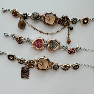 Pode incluir: Três pulseiras de relógio decorativas com vários berloques e enfeites. Os relógios têm mostradores quadrados e em forma de coração com metal dourado e prateado. As pulseiras apresentam berloques decorativos, incluindo botões, estrelas e desenhos florais.
