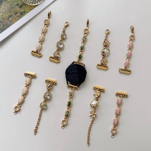 Peut inclure: Une collection de bracelets de montre et de breloques décoratives en métal doré, ornés de diverses pierres incrustées, notamment des cabochons ovales crème, roses et verts, des cristaux facettés transparents et des motifs en forme de cœur.