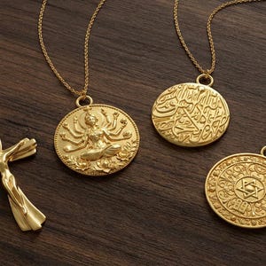 Puede incluir: Collares dorados con colgantes religiosos y espirituales. La colección incluye una cruz, un colgante con una deidad, un colgante con escritura árabe y un colgante con un diseño de estrella. Los collares se exhiben sobre una superficie de madera oscura.