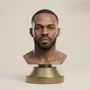 May include: A sculpted bust of a man with dark hair and a beard. The bust is a light brown colour and is mounted on a gold-coloured base. The sculpture is a detailed representation of a person's head and shoulders.