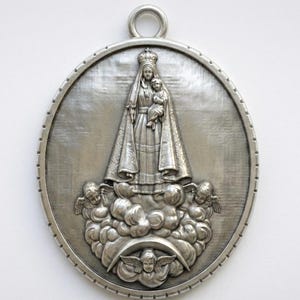 Puede incluir: Colgante ovalado plateado con un relieve detallado de una figura religiosa sosteniendo a un niño, rodeada de nubes y ángeles. El colgante tiene un lazo en la parte superior para colgar.