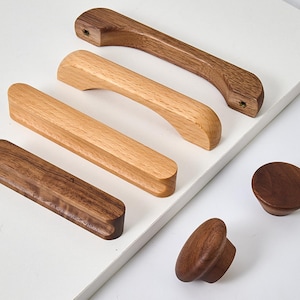 Puede incluir: Una colección de herrajes de madera para muebles que incluye tres tiradores de cajón alargados en varios tonos de madera y dos pomos circulares. Los tiradores presentan diseños suaves y curvos con vetas de madera visibles y tonos oscuros.