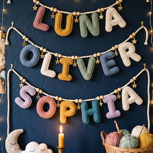 May include: Three horizontal garlands featuring crocheted letters spelling LUNA, OLIVER, and SOPHIA. The letters are in various colors including pink, yellow, green, blue, and cream, accented with wooden beads and small gold star charms on white rope.
