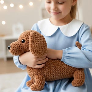 May include: A brown crocheted dachshund dog toy with black eyes and floppy ears. The handmade stuffed animal is held by a person wearing a blue dress with a white collar. The toy is a soft, cuddly item.