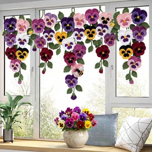 Può includere: Una tendina a uncinetto con viole colorate nei toni del viola, rosa, giallo e bianco. I fiori sono attaccati a viti verdi, creando un design floreale decorativo. Un vaso di viole si trova sul davanzale.