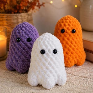 May include: Three plush ghost figures crafted from soft, textured yarn. One ghost is purple, one is white, and one is orange. Each figure features two small, round, black plastic eyes positioned on the front, with a rounded top and a scalloped base.