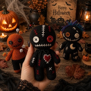 May include: A black crocheted voodoo doll with a red heart patch, white and red button eyes, and white stitched details. The doll features pins in the head and is held in front of two other crocheted dolls, a pumpkin, and a Happy Halloween sign.
