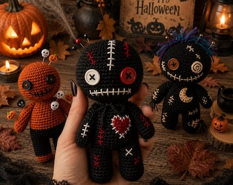 Puede incluir: Una muñeca vudú de ganchillo negra con un parche de corazón rojo, ojos de botón blancos y rojos y detalles cosidos en blanco. La muñeca tiene alfileres en la cabeza y se sostiene frente a otras dos muñecas, una calabaza y un cartel de Halloween.