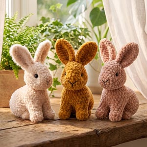 May include: Three plush rabbit toys made of soft, textured yarn. The rabbit on the left is cream-colored, the center rabbit is mustard yellow, and the rabbit on the right is dusty rose. Each rabbit features long upright ears and dark eyes.
