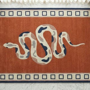 Könnte beinhalten: Ein rechteckiger Teppich mit einer rostorangen Mitte und einem großen, geschlängelten Schlangenmotiv in Creme-, Beige- und Dunkelblautönen. Der Teppich ist von einem cremefarbenen Rand mit einem wiederkehrenden Muster aus dunkelblauen Quadraten umgeben.