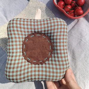May include: A square, padded potholder with a brown and teal gingham pattern. A circular brown patch is sewn onto the center with teal stitching. A cream-colored fabric loop is attached to the top for hanging. The potholder is designed for kitchen use.