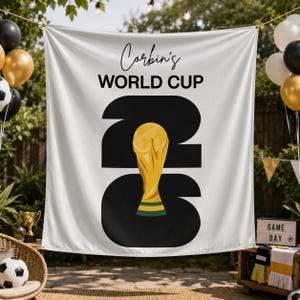 May include: A white fabric banner featuring the black text Corbin's WORLD CUP above large black numbers 2026. A gold-colored trophy illustration replaces the zero in the year. The banner is displayed outdoors with black and gold balloons.