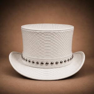 Puede incluir: Un sombrero de copa blanco con un patrón texturizado de piel de serpiente en la copa y el ala. El sombrero está acentuado con una banda blanca a juego en la base, decorada con una fila de tachuelas de metal plateado pulido espaciadas.