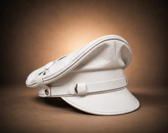 May include: A white leather military-style peaked cap featuring a structured crown, a stiff visor, and a horizontal band across the front. The band is secured with a small silver-toned metal button on the side, providing a clean and uniform appearance.