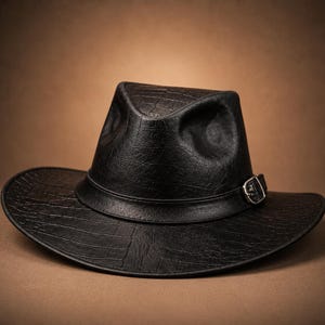 Peut inclure: Un chapeau fedora noir avec un motif texturé en peau de crocodile sur la calotte et les bords. Le chapeau comprend une bande noire assortie autour de la base de la calotte, agrémentée d'une petite boucle en métal argenté sur le côté.