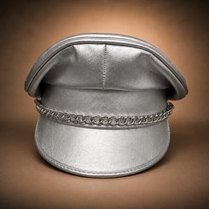 May include: A metallic silver military-style cap featuring a structured crown and a stiff, curved visor. A polished silver-tone metal chain is attached across the front of the cap, resting just above the brim, adding a decorative element to the design.