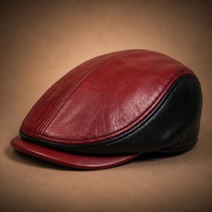 May include: A flat cap constructed from leather with a smooth, deep red top panel and a textured black side panel. The cap features a short, stiff red brim and visible stitched seams that define the rounded shape of the headwear piece.