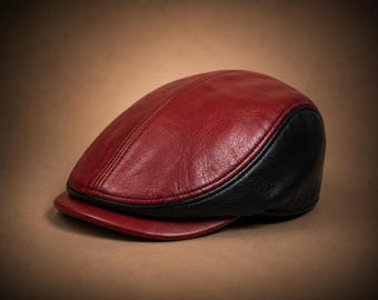 May include: A flat cap constructed from leather with a smooth, deep red top panel and a textured black side panel. The cap features a short, stiff red brim and visible stitched seams that define the rounded shape of the headwear piece.