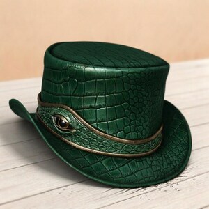 May include: A dark green top hat featuring a textured crocodile skin pattern. The hat is encircled by a matching green band with a metallic gold trim and a central decorative eye element with a brown iris and a black pupil centered on the front.
