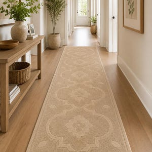 Puede incluir: Una alfombra larga y rectangular de color beige con un estampado floral, colocada en un pasillo con suelo de madera clara. El diseño de la alfombra presenta motivos florales estilizados repetidos. El pasillo tiene una paleta de colores neutros, con una consola de madera y plantas en macetas.
