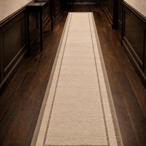 Puede incluir: Una alfombra rectangular beige para pasillo con un borde doble en un tono beige más oscuro. La alfombra está colocada sobre un suelo de madera oscura. La alfombra es de forma larga y estrecha, adecuada para pasillos.