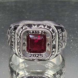 May include: Silver class ring featuring a square, deep red gemstone. The ring is inscribed with "A VAL" and "CLASS OF 2025". The band has detailed patterns. A graduation ring.