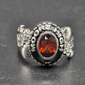 May include: A silver ring featuring a large, oval, red gemstone. The ring is inscribed with the words "MORE HIGH". The ring has floral detailing on the sides. The ring is a piece of jewellery.