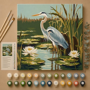 May include: A paint-by-numbers kit featuring a blue and white heron standing in a pond with white water lilies and tall reeds. The set includes a canvas, three paintbrushes, and twenty-four numbered paint pots with a color reference guide.