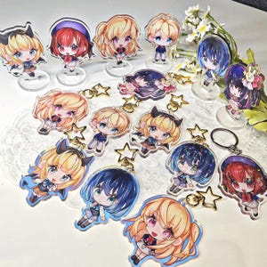 Może przedstawiać: Kolekcja akrylowych figurek i breloków w stylu anime z postaciami chibi o żywych kolorach włosów, w tym blond, czerwonym i niebieskim. Zestaw zawiera okrągłe podstawki do ekspozycji oraz złote kółka na klucze w kształcie gwiazdek.