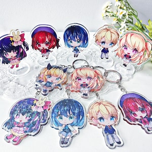 May include: A selection of anime-style acrylic charms and keyrings featuring colourful chibi characters. The charms are on clear stands, while the keyrings have silver rings. The characters have various hair colours and outfits.