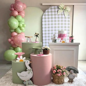 May include: A whimsical party scene with pastel pink and green balloons, pedestals, and decorations. A cake, flowers, and duck figurines are displayed. The backdrop features a gingham pattern and a large bow.