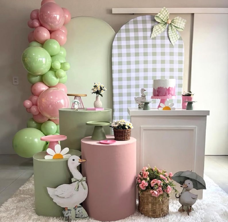 May include: A whimsical party scene with pastel pink and green balloons, pedestals, and decorations. A cake, flowers, and duck figurines are displayed. The backdrop features a gingham pattern and a large bow.