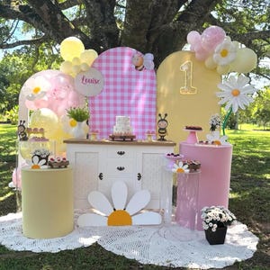 May include: A whimsical outdoor party setup with a daisy theme. Features include a white cabinet, pink and yellow pedestals, a cake, balloons, and a large daisy cutout. The backdrop includes a pink gingham pattern and the number "1".