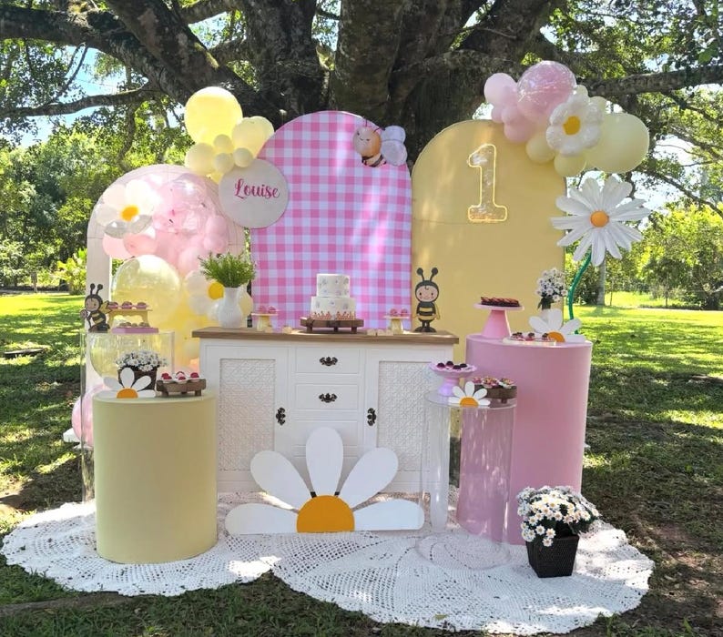 May include: A whimsical outdoor party setup with a daisy theme. Features include a white cabinet, pink and yellow pedestals, a cake, balloons, and a large daisy cutout. The backdrop includes a pink gingham pattern and the number "1".