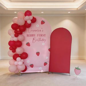 May include: A pink backdrop with the text "Veronica's BERRY FIRST Birthday" and strawberry illustrations. A cluster of red and pink balloons is on the left. A red arch-shaped structure is on the right.
