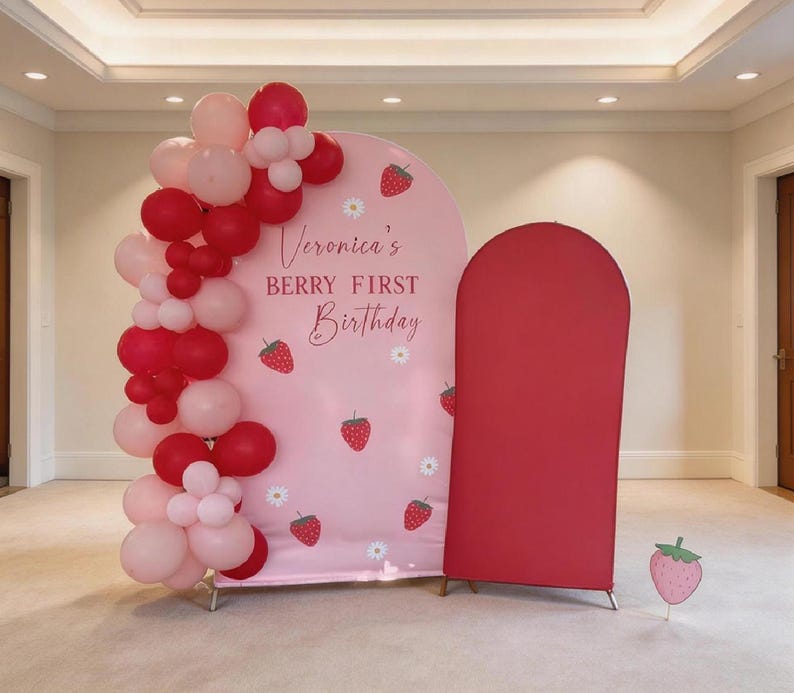 May include: A pink backdrop with the text "Veronica's BERRY FIRST Birthday" and strawberry illustrations. A cluster of red and pink balloons is on the left. A red arch-shaped structure is on the right.