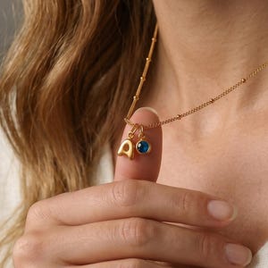 May include: Gold-colored necklace with a delicate chain and two charms. One charm is the letter "A", and the other is a small, round, blue gemstone. The necklace is worn against a cream-colored sweater.