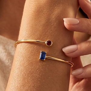 May include: A gold-toned open cuff bracelet featuring a round, deep red gemstone at one end and a rectangular, vibrant blue gemstone at the other. The metal band is smooth and polished, creating a minimalist design that rests on the wrist.