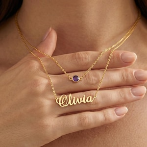 Puede incluir: Un conjunto de collar dorado de dos hilos. El hilo superior presenta una pequeña gema redonda de color púrpura. El hilo inferior deletrea el nombre "Olivia" en escritura cursiva. El collar se muestra en una mano.